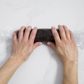 two hands rolling dark dough wrapped in plastic into a cylinder